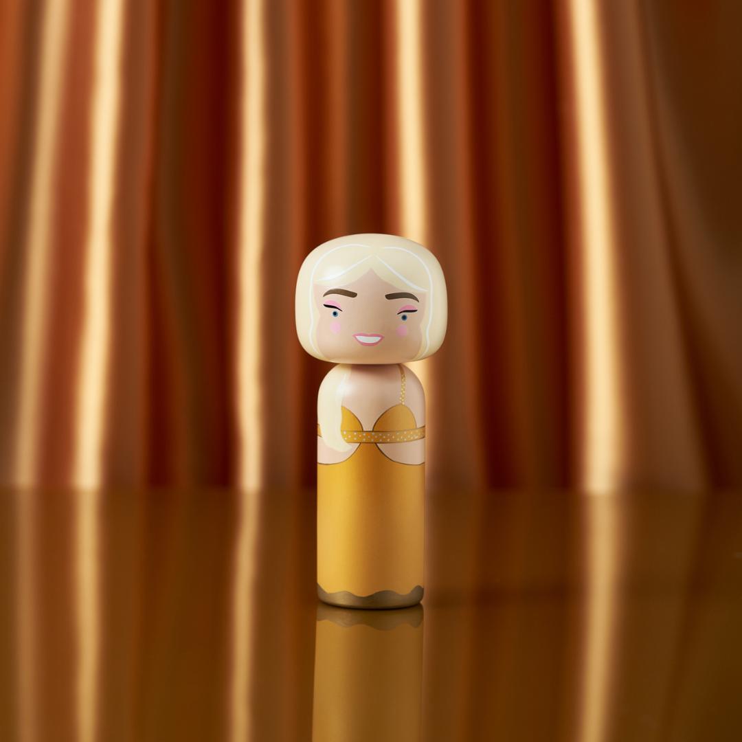 Une poupée kokeshi avec une robe jaune et des cheveux blonds se tient sur un fond orange rayé et brillant.