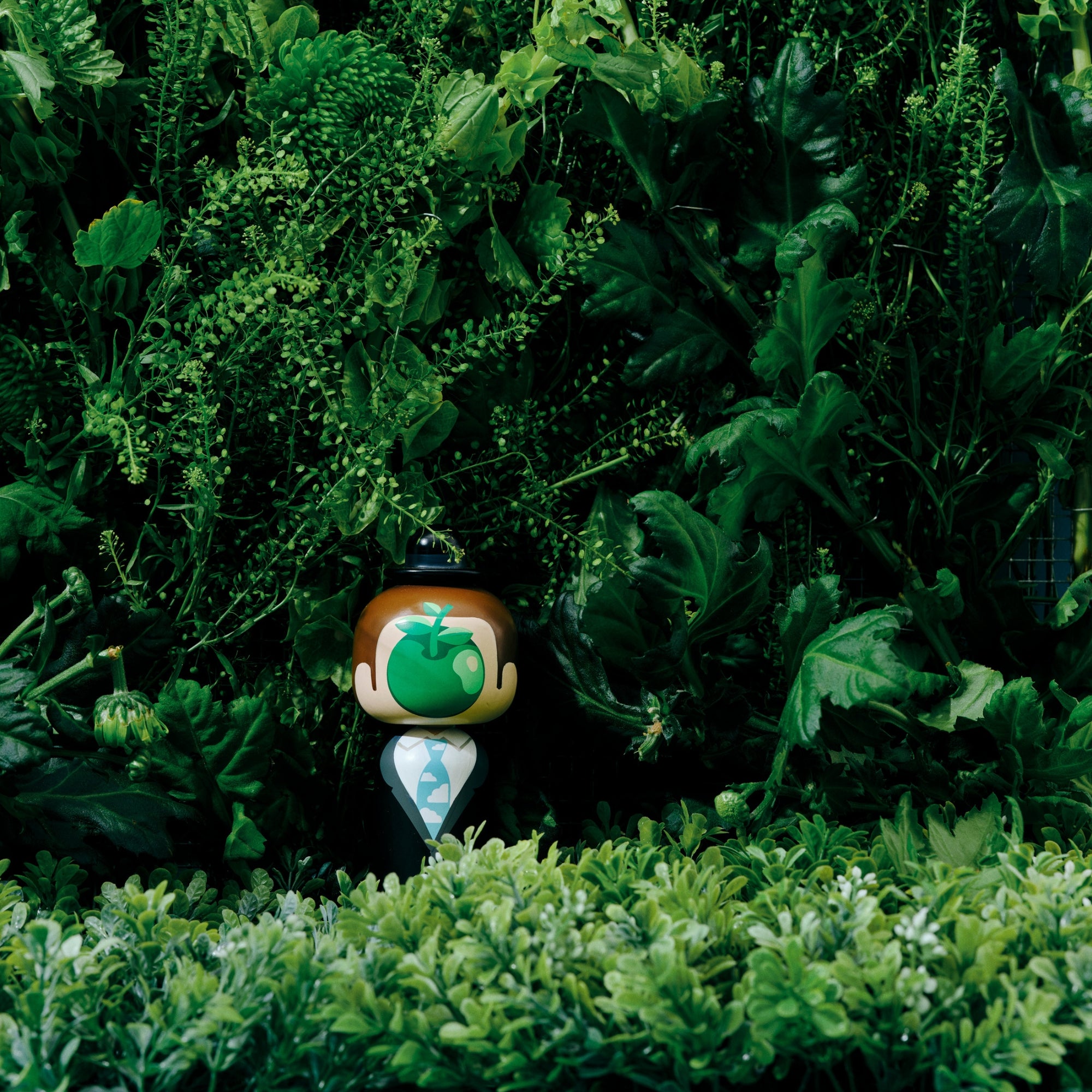 Une poupée kokeshi avec un visage en forme de pomme verte se tient au milieu d'un feuillage vert luxuriant.
