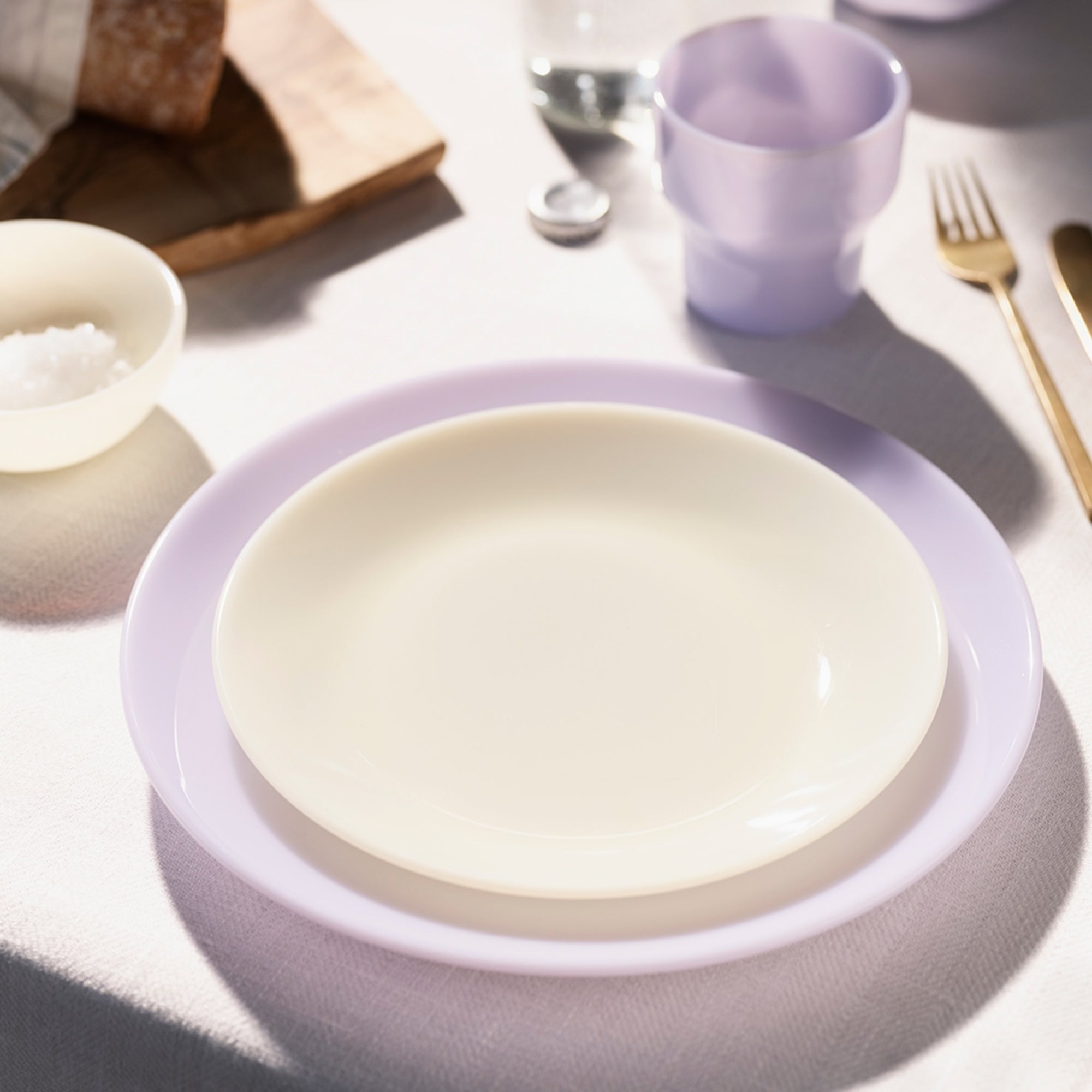 Une table avec une assiette blanche sur une assiette lavande, à côté d'une tasse violette et de couverts dorés.