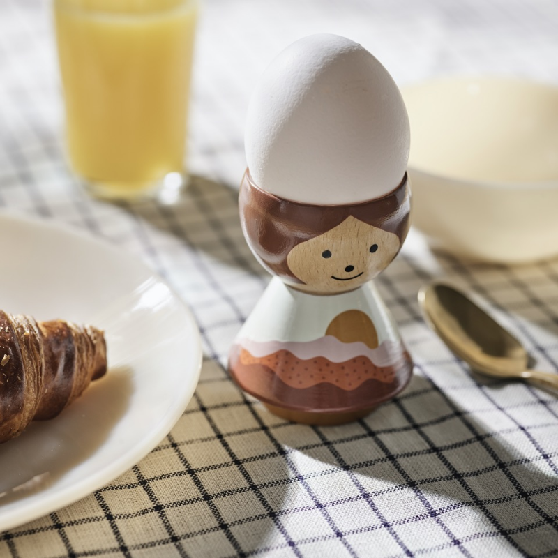 Coquetier de poupée Kokeshi avec un visage souriant, tenant un œuf, sur une nappe à carreaux avec des articles de petit déjeuner.