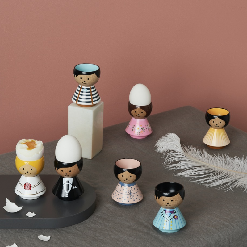 Coquetiers de poupées Kokeshi aux motifs et couleurs variés sur une table à fond rose avec une plume blanche.