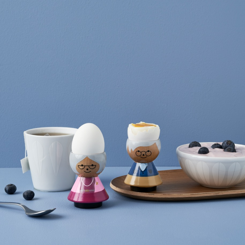 Deux coquetiers kokeshi avec des œufs, une tasse de thé et un bol de yaourt avec des myrtilles sur un plateau en bois.