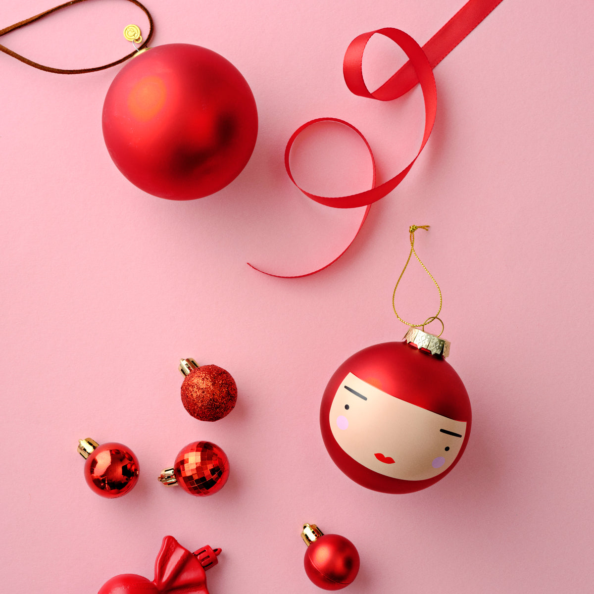 Red Christmas ornaments, including a kokeshi doll face, on a pink background with a red ribbon.