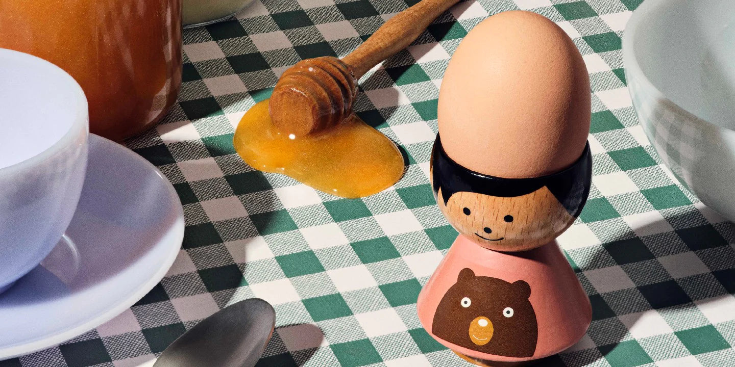 Coquetier de poupée Kokeshi avec un motif d'ours sur une nappe à carreaux, entouré de miel et de plats.