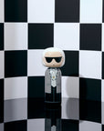 Une poupée kokeshi portant des lunettes de soleil et un costume se détache sur un fond à carreaux noirs et blancs.