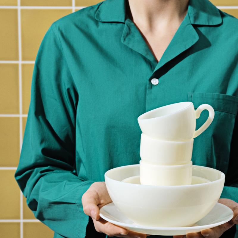 Personne en chemise verte tenant des tasses et des bols blancs empilés sur un fond carrelé jaune.