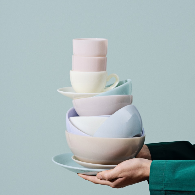 Mains tenant une pile de tasses et de bols aux couleurs pastel sur un fond bleu clair.