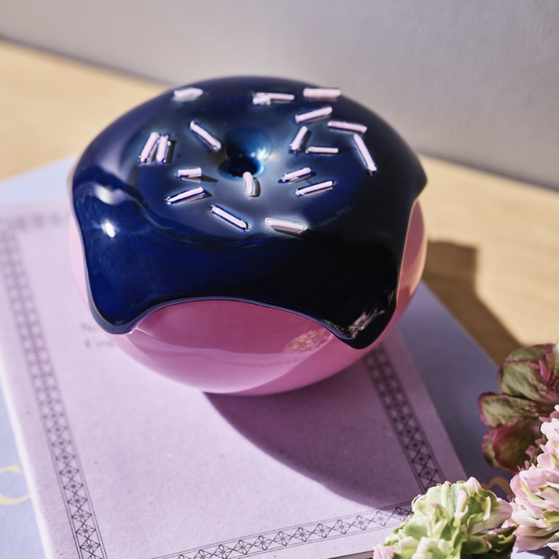 Un beignet en céramique rose et bleu avec des éclats sur un livre violet, à côté de fleurs.