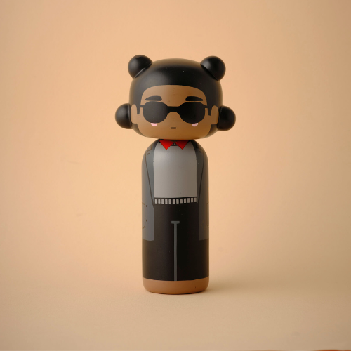 Une poupée kokeshi avec des lunettes de soleil, des cheveux noirs et une tenue grise sur un fond beige.