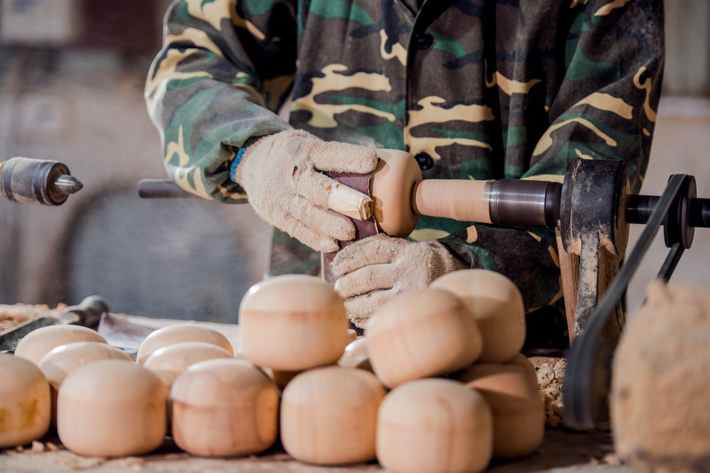 Personne en veste de camouflage fabriquant des poupées en bois sur un tour, avec des pièces finies au premier plan.