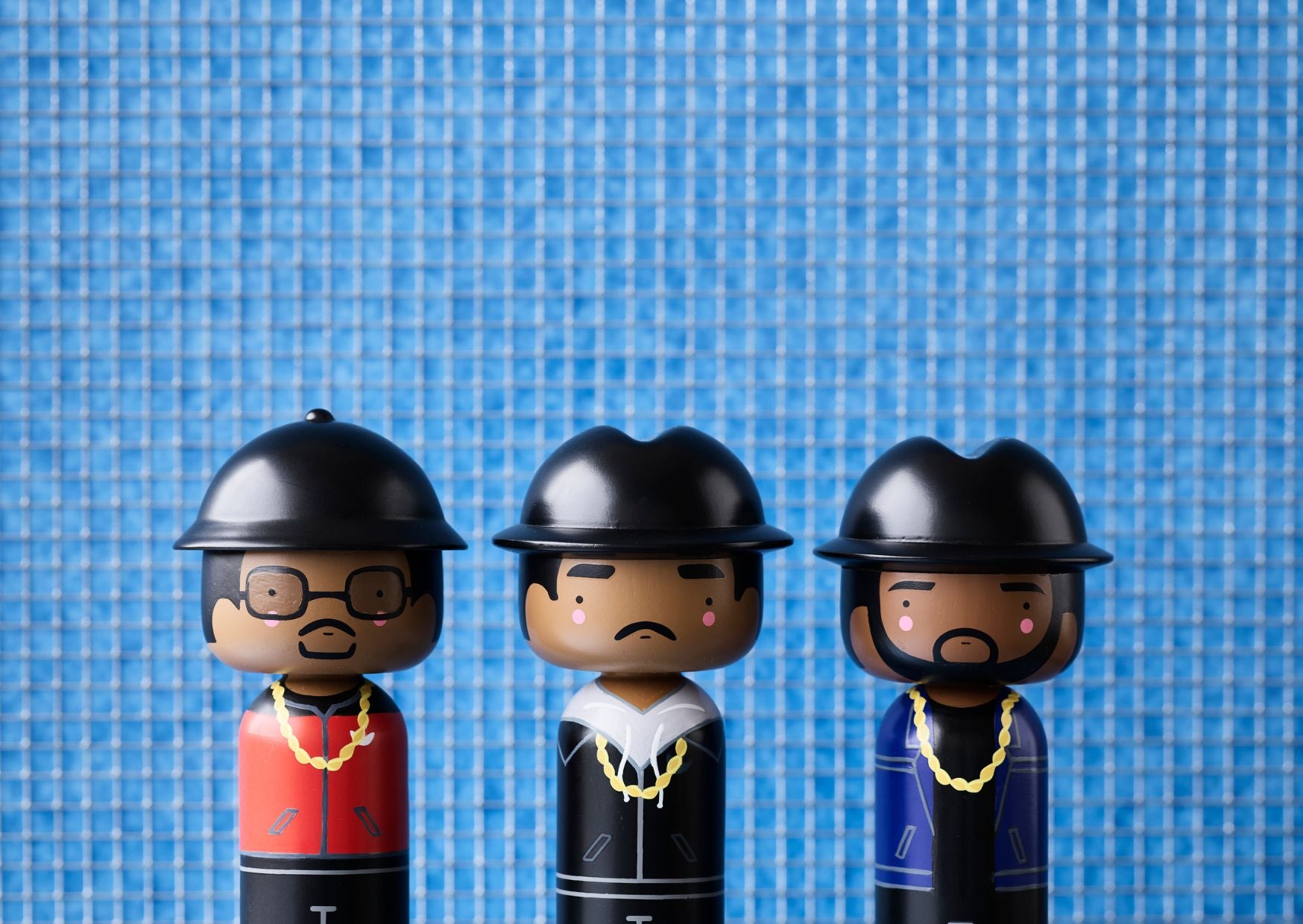 Trois poupées kokeshi avec des chapeaux et des chaînes sur un fond carrelé bleu.
