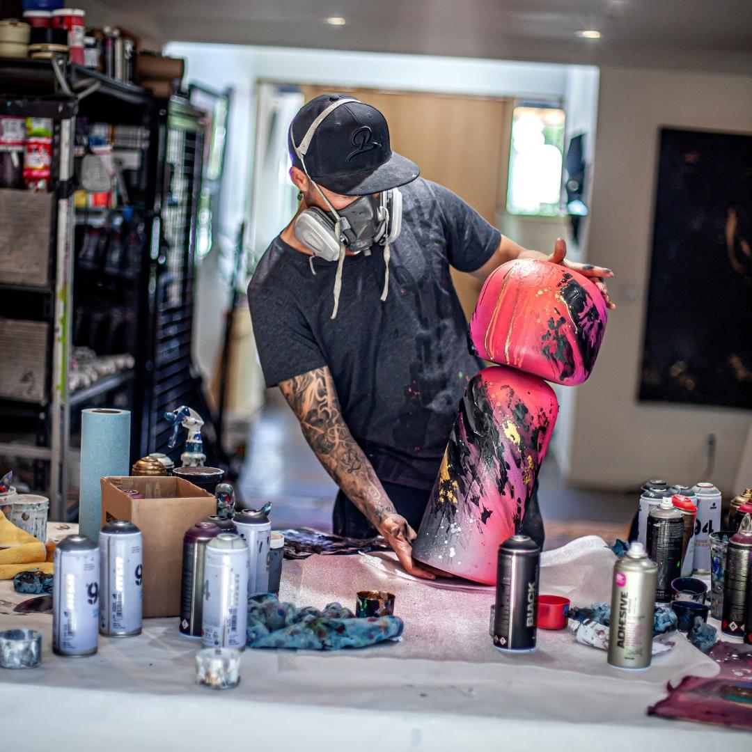 En kunstner iført maske maler store kokeshi-dukker i et studio fylt med spraymalingsbokser.