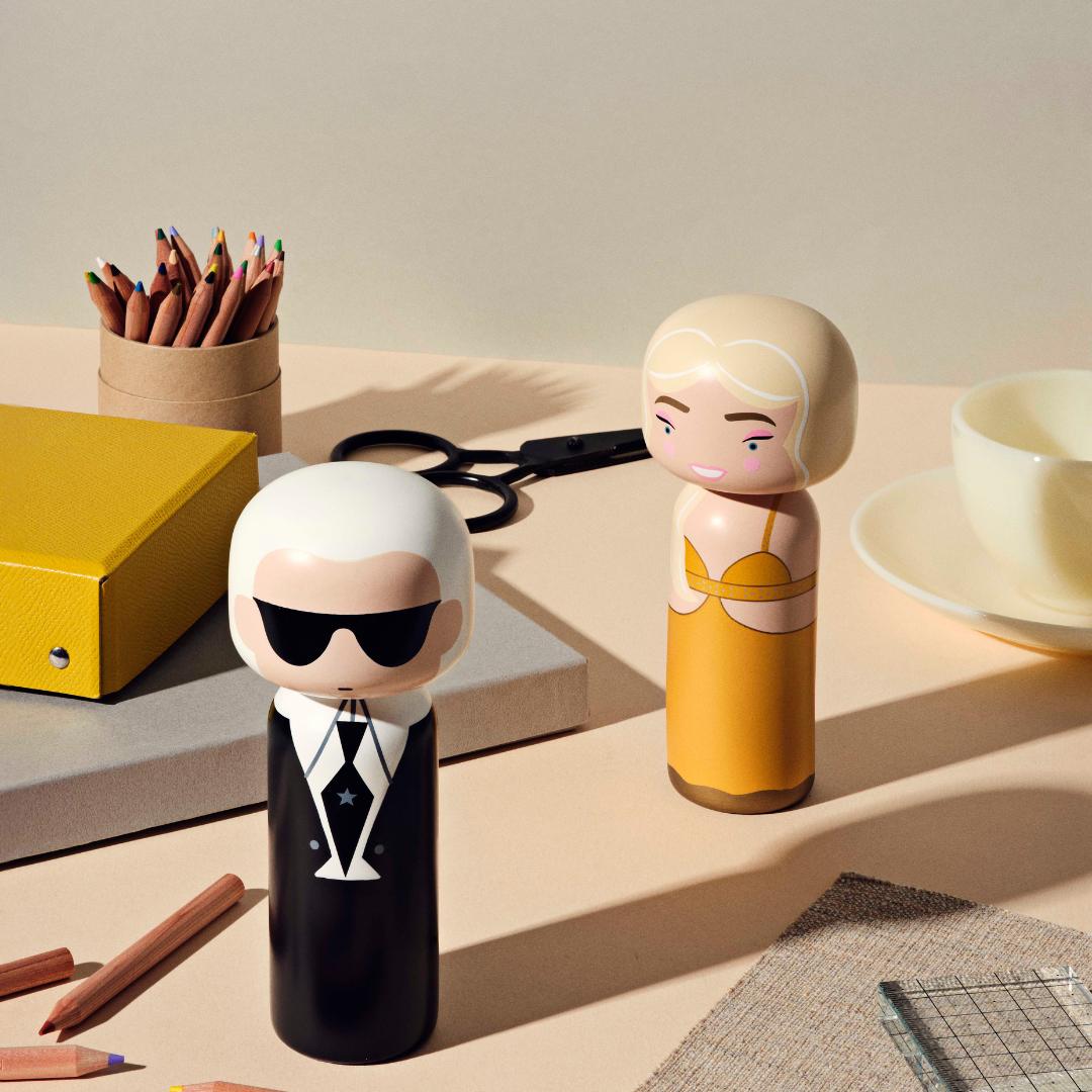 Kokeshi-dukker som ligner kjente figurer på et skrivebord med blyanter, en gul perm og en kopp.