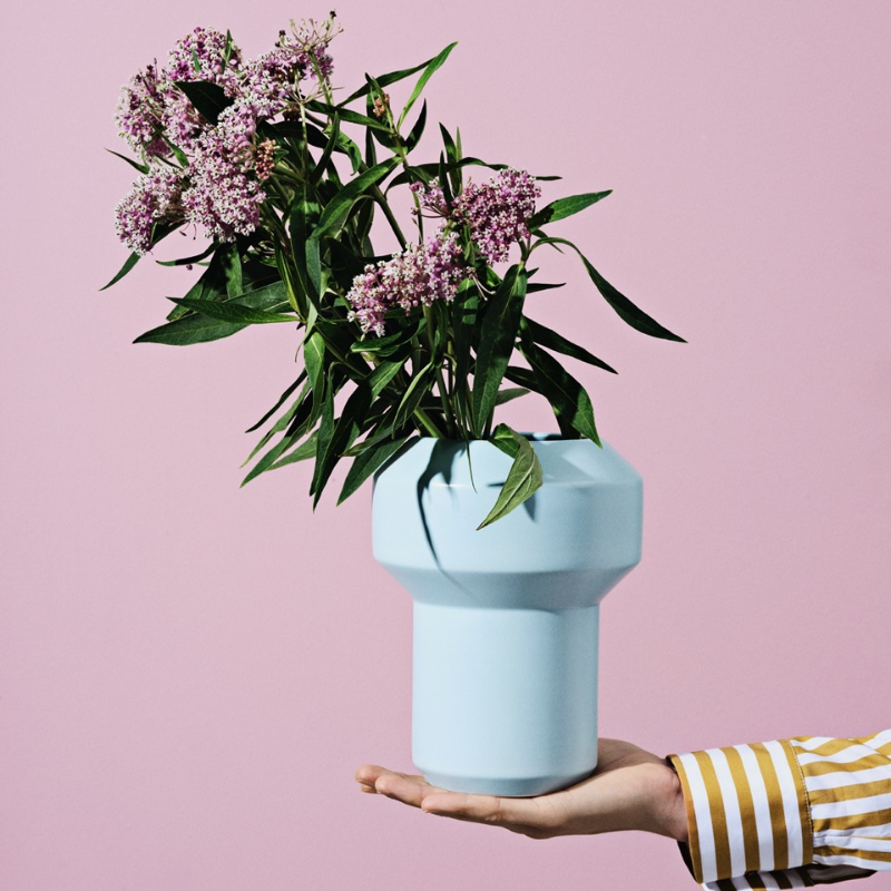 En hånd holder en lyseblå geometrisk vase med rosa blomster mot en rosa bakgrunn.
