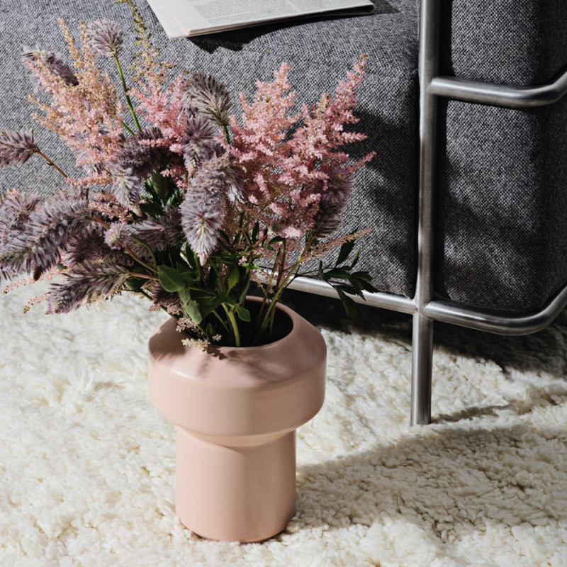 En rosa vase med rosa og lilla blomster på et hvitt teksturert teppe ved siden av en grå sofa.