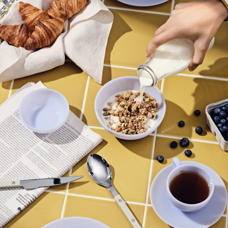 En frokostscene med frokostblanding, milk , croissanter, blåbær, te og en avis på et gult flislagt bord.