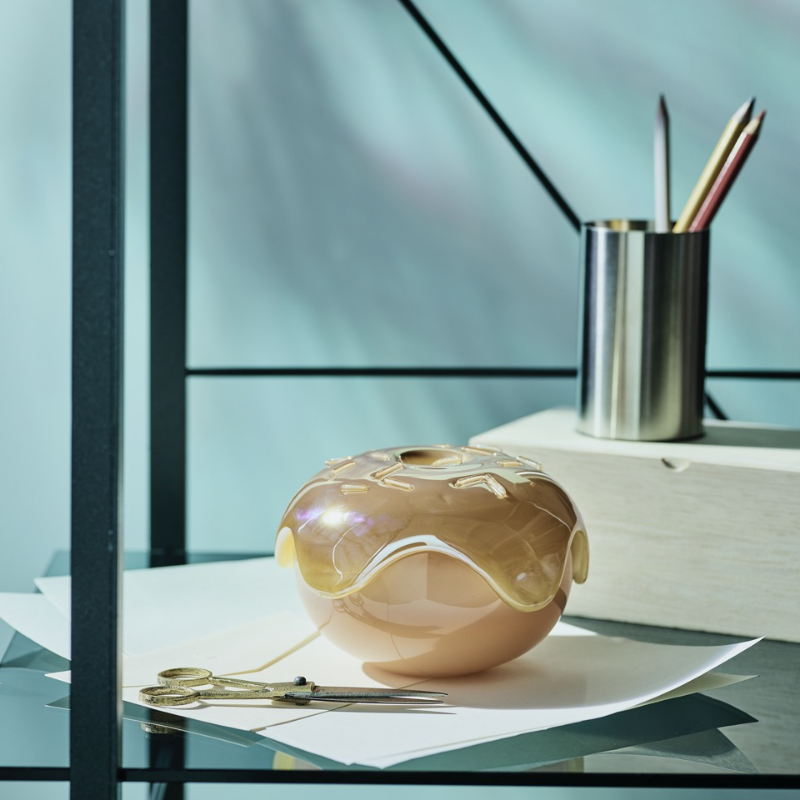 En blank, smultringformet vase med karamellglasurdesign på en glasshylle, ved siden av saks og blyanter.