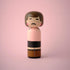 Lucie Kaas wooden kokeshi doll with brown hair, mustache, pink cheeks and black belt on pink background