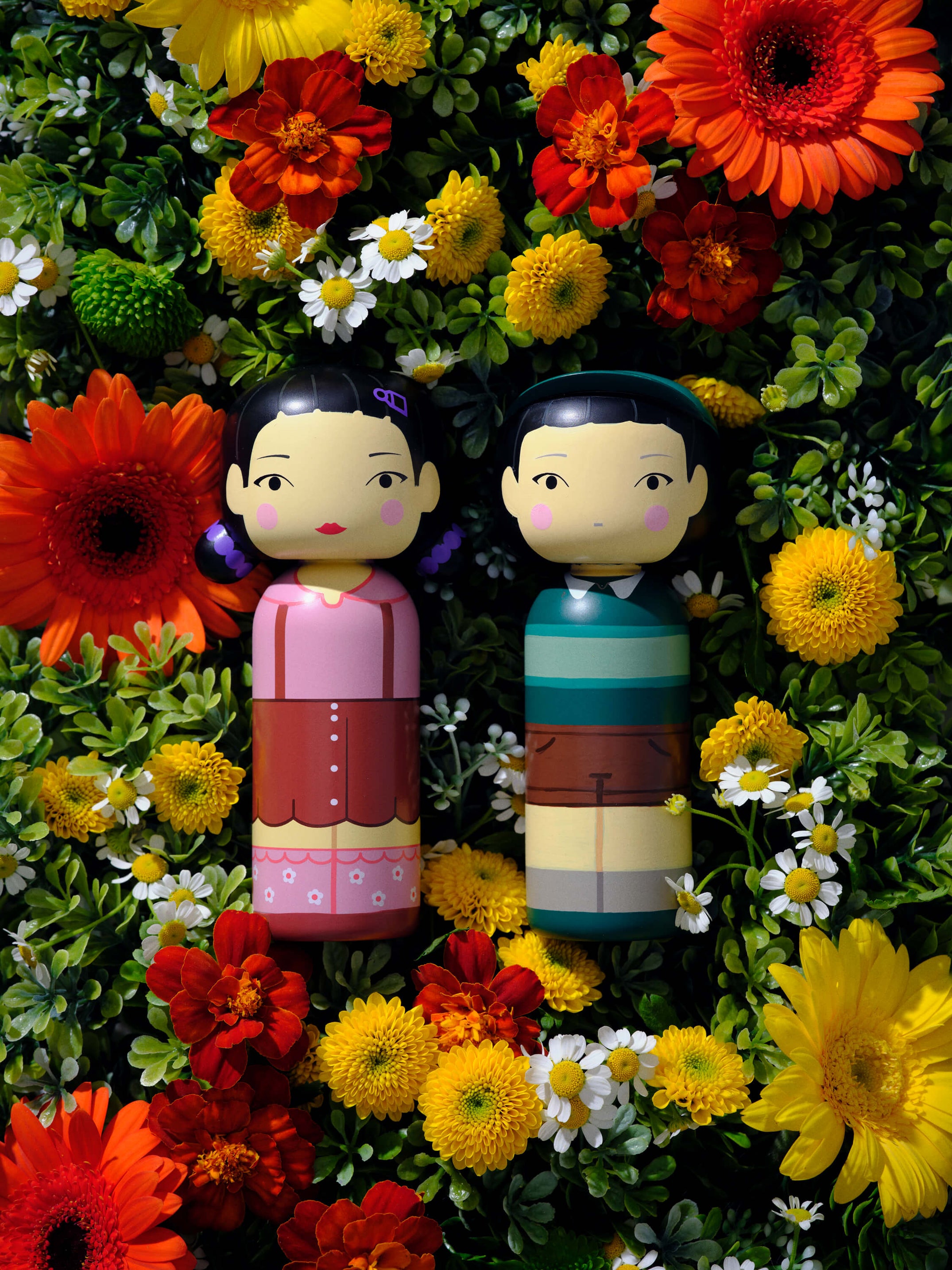 Zwei Kokeshi-Puppen auf einem Bett aus bunten Blumen, darunter Gänseblümchen und Gerbera.