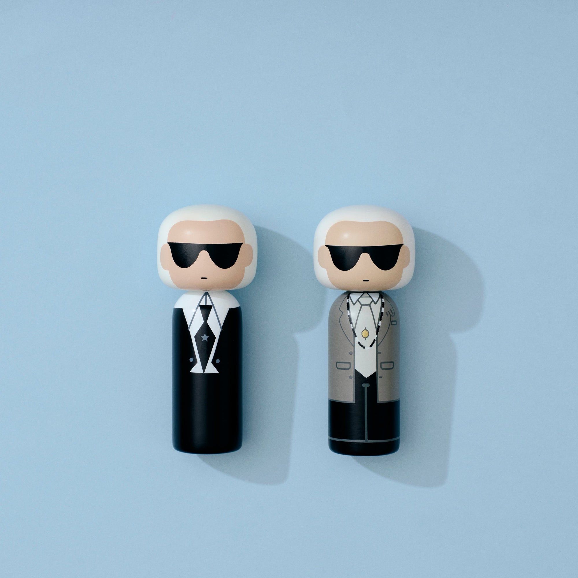 Zwei Kokeshi-Puppen mit Sonnenbrillen und stilvollen Outfits auf einem hellblauen Hintergrund.