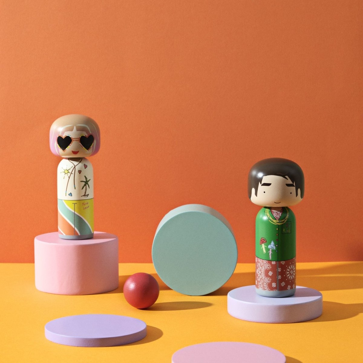 Mira Mikati und Gio Lucie Kaas Kokeshi-Puppen aus der Mira Mikati-Kollektion in einem orangefarbenen Rahmen mit Dekorationen