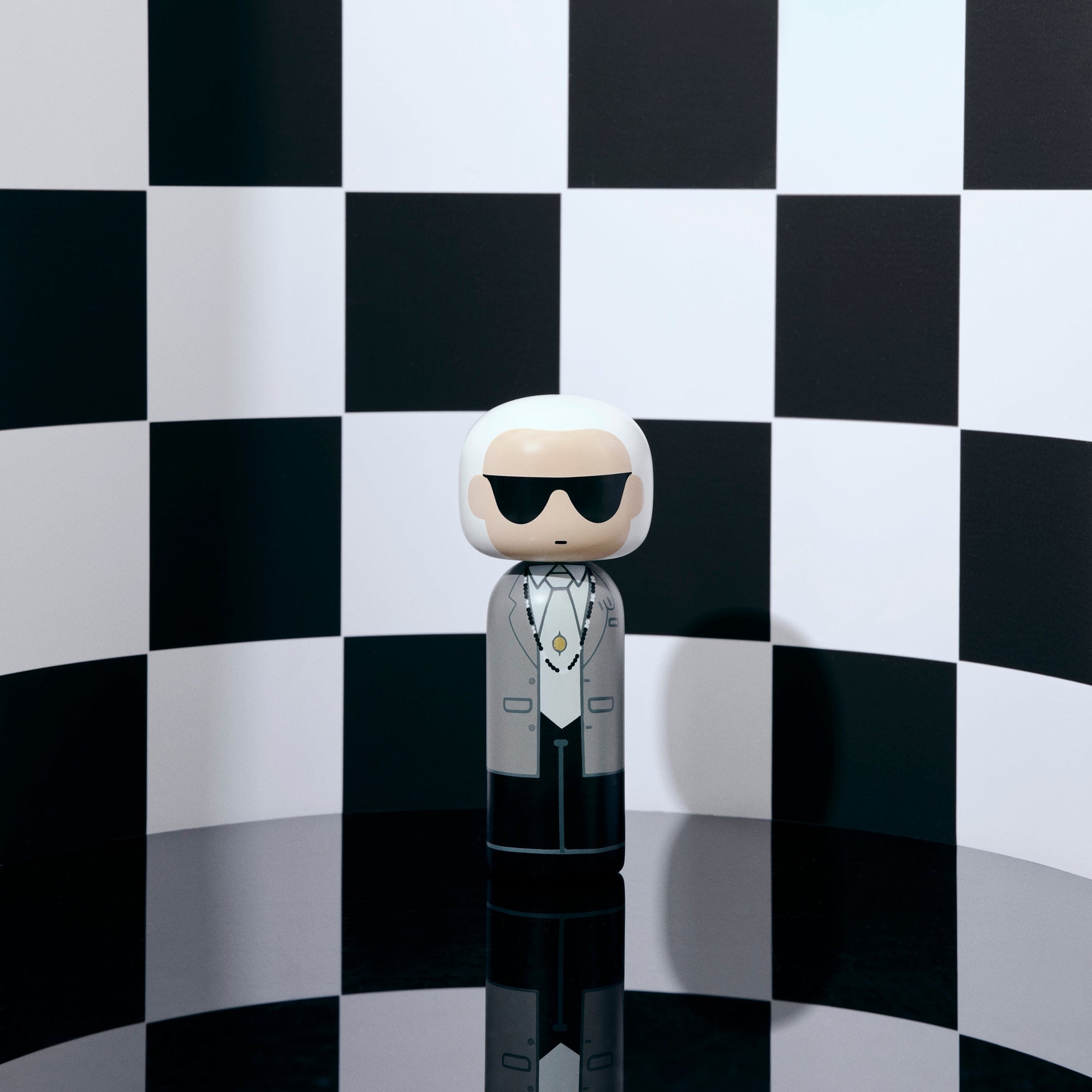 Une poupée kokeshi portant des lunettes de soleil et un costume se détache sur un fond à carreaux noirs et blancs.