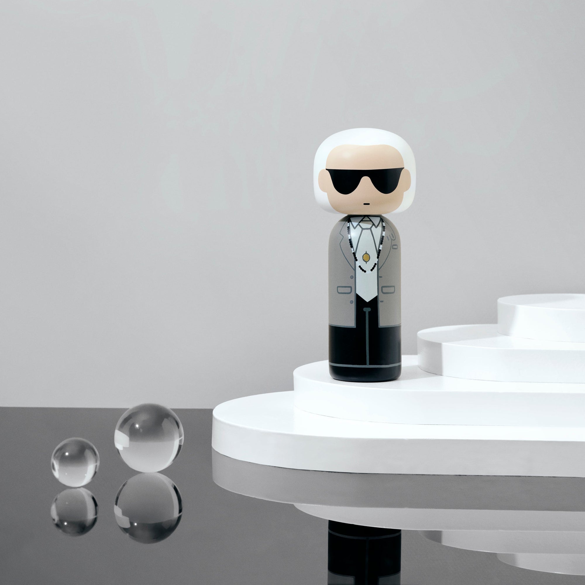 Poupée Kokeshi avec des lunettes de soleil sur une plate-forme blanche, avec des sphères réfléchissantes à proximité.