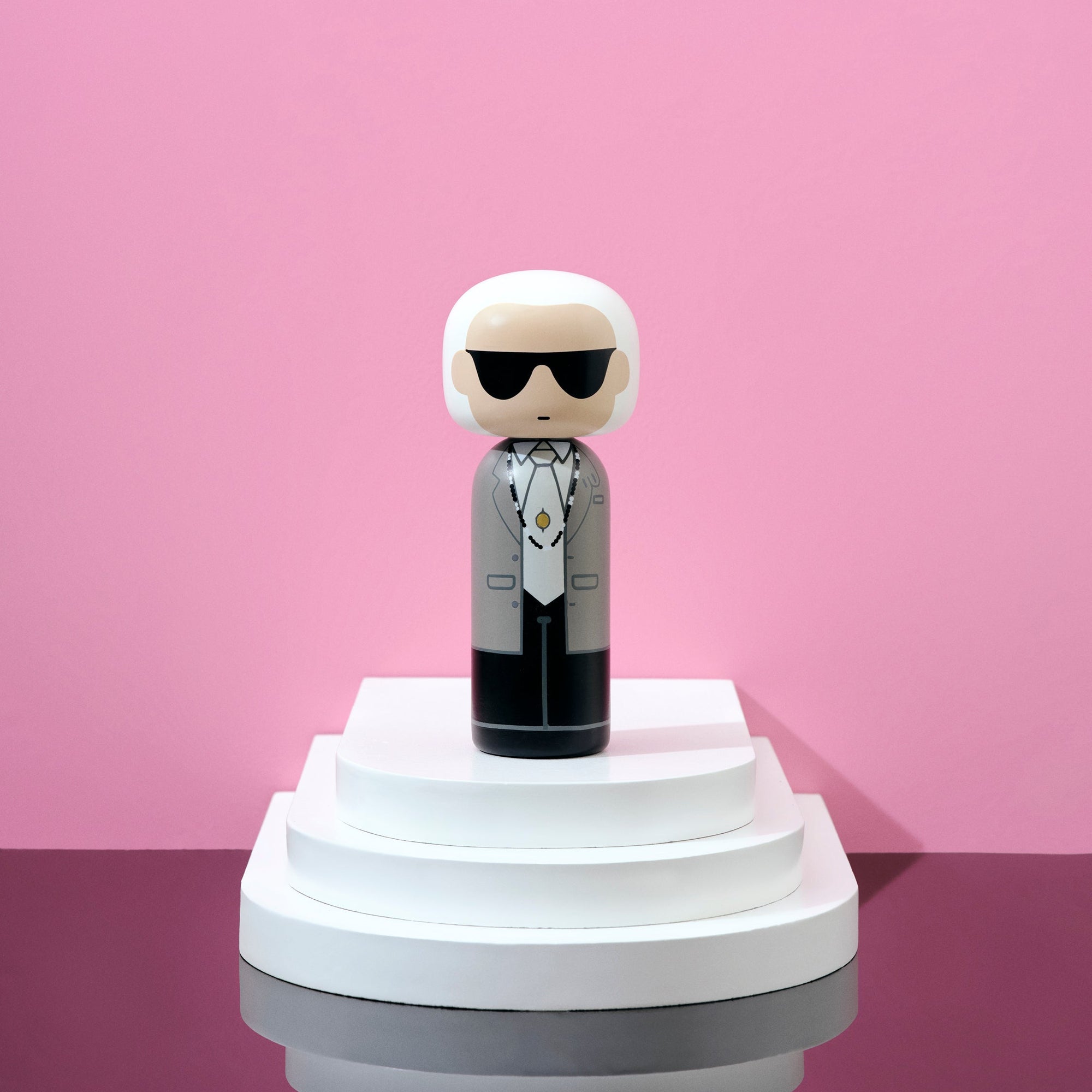 Poupée Kokeshi portant des lunettes de soleil et un costume sur une plate-forme blanche à étages sur fond rose.